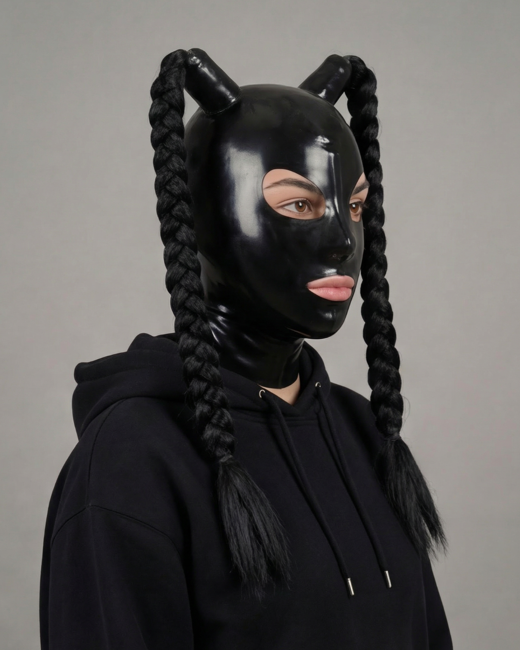Person wearing a black latex hood with braided hair extensions on a gray background