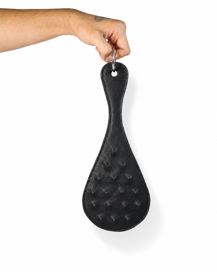 Trapunto Paddle held by O-ring loop, back view showing smooth black leather and raised trapunto pattern, hand model with tattoo, Leathersubculture