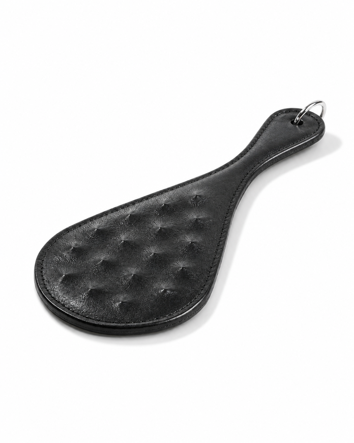 Trapunto Paddle flat lay angled — black leather BDSM impact paddle with raised trapunto diamond stud pattern on face, O-ring at handle, handcrafted by Leathersubculture