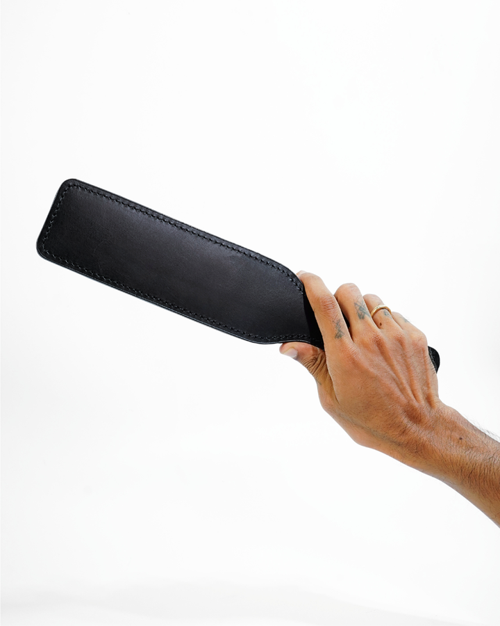 Genuine leather spanking paddle from BDSM Bondage Kit - smooth black leather with stitched edges, shown held in hand, handcrafted by Leathersubculture
