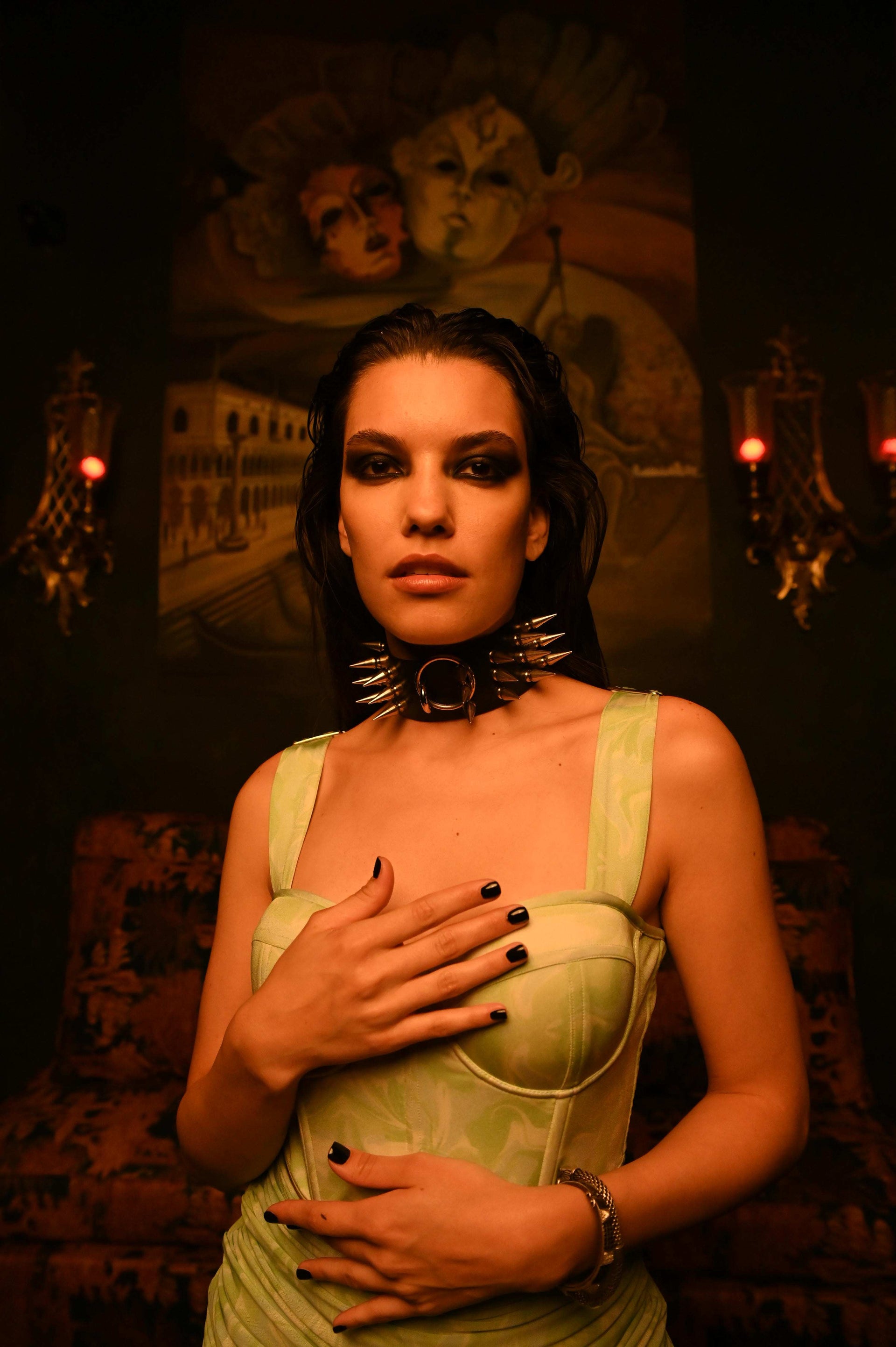 Woman in a green dress with dark makeup and jewelry in a dimly lit room.